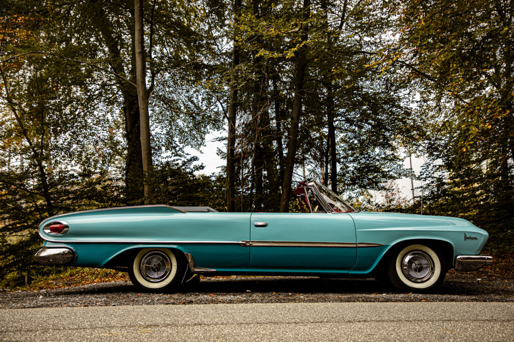 Zijaanzicht van een elegant lichtblauwe klassieke Dodge Polara cabriolet, met open dak, geparkeerd aan de rand van een bos met hoge bomen.