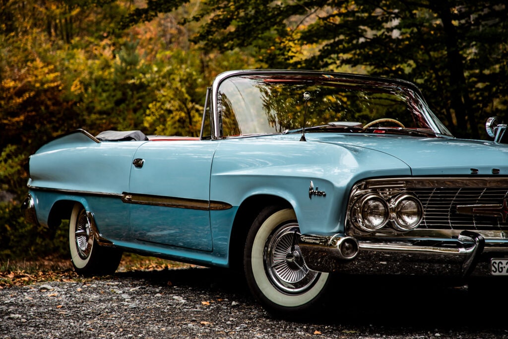 Elegante Klassieke Dodge Polara Cabriolet in Herfstbos Zijaanzicht van een lichtblauwe klassieke Dodge Polara cabriolet met open dak en whitewall banden, geparkeerd op een grindpad met herfstbomen op de achtergrond.