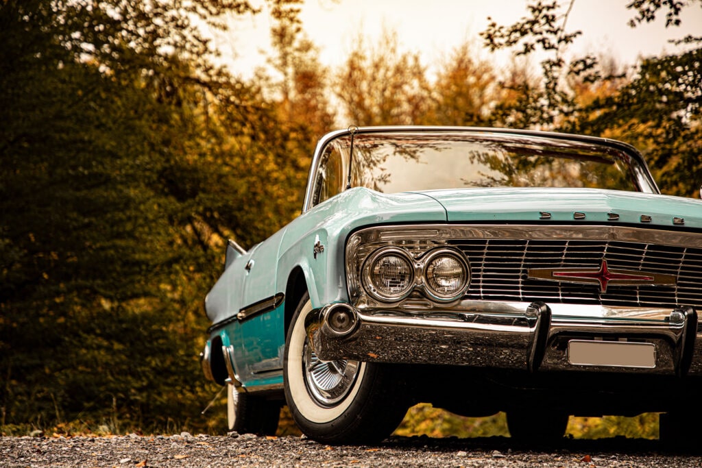Laag vooraanzicht van een lichtblauwe klassieke Dodge Polara cabriolet, geparkeerd op een grindpad, met herfstbladeren op de achtergrond.
