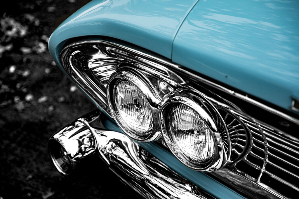 Close-up van de dubbele chromen koplampen en de grille van een lichtblauwe klassieke Dodge Polara, met een onscherpe zwart-witte achtergrond.