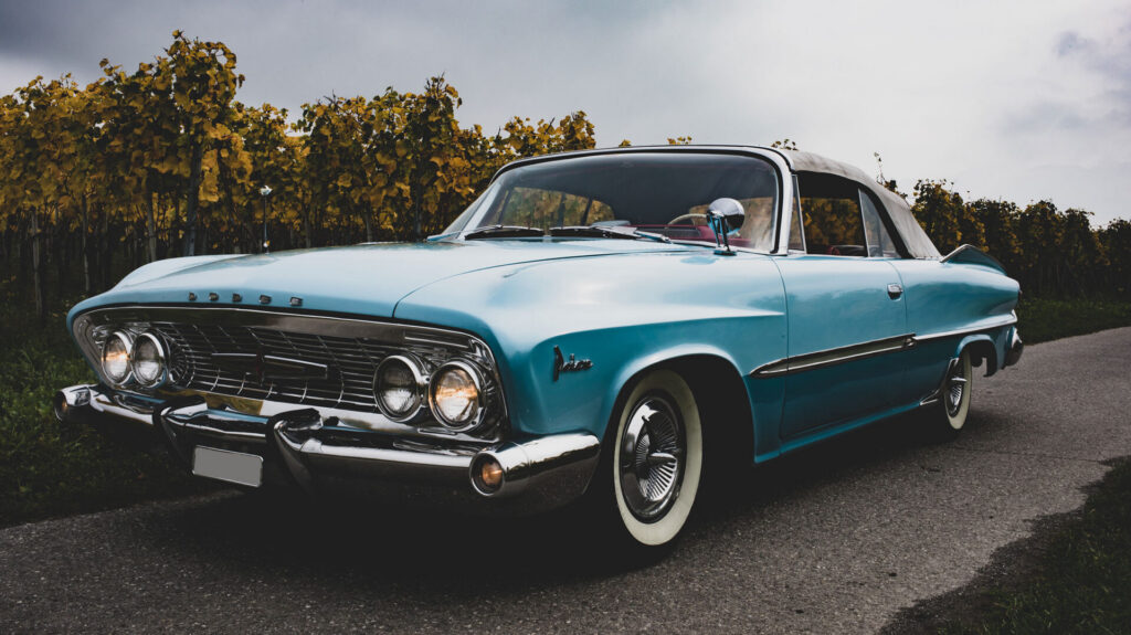 Een lichtblauwe klassieke Dodge Polara cabriolet, geparkeerd op een geasfalteerde landweg met een wijngaard in herfstkleuren op de achtergrond.