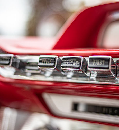 Close-up van de chromen drukknopbediening voor de automatische transmissie (P, R, N, DRIVE, SECOND, FIRST) op het rode dashboard van een klassieke auto.