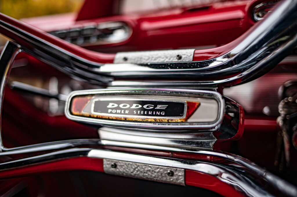 Close-up van het chromen midden van een klassiek stuurwiel, met daarop "DODGE POWER STEERING" en een rode achtergrond, tegen een onscherp rood interieur.