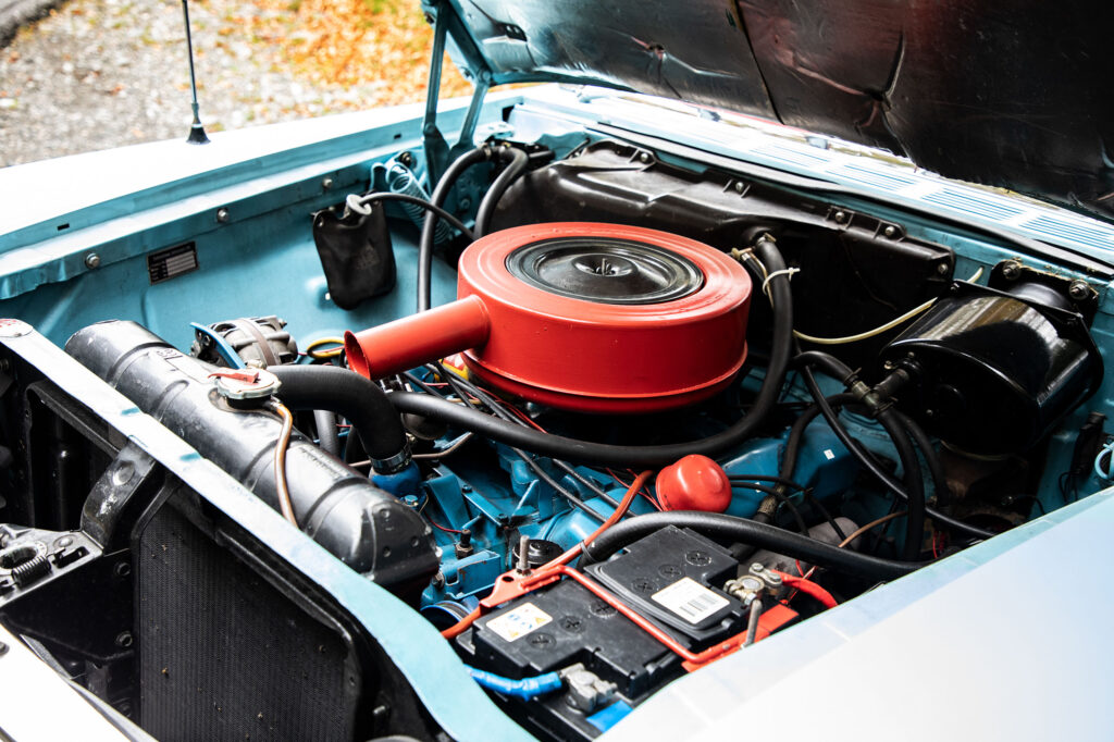 Close-up van de open motorkap van een lichtblauwe klassieke auto, die een gedetailleerde weergave van de motorruimte met rode luchtfilterbehuizing en diverse motoronderdelen toont.