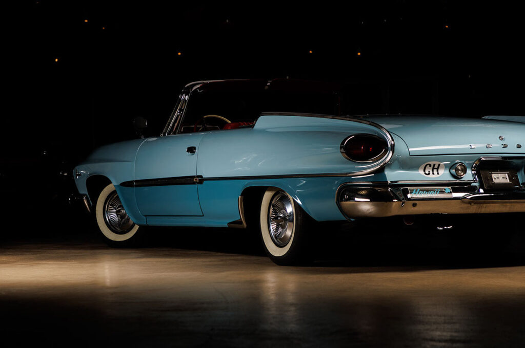 Achteraanzicht van een lichtblauwe klassieke Dodge Polara cabriolet, verlicht door een spotlicht in een donkere, minimalistische ruimte, met reflecties op de vloer.